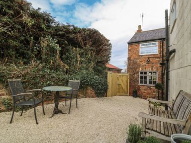 A small outdoor courtyard with gravel ground two black chairs and a round table next to a hedge and a wooden bench beside a brick building at New Inn Apartment in Tholthorpe near Easingwold