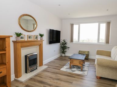 A living room with a wooden fireplace mantel, wall-mounted TV, beige sofa, wooden coffee table, indoor plants, and large window at The Croft Bungalow in Plumbland near Aspatria