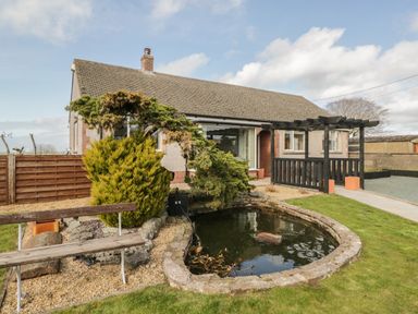 A bungalow with a pond and a wooden bench in the garden at The Croft Bungalow in Plumbland near Aspatria