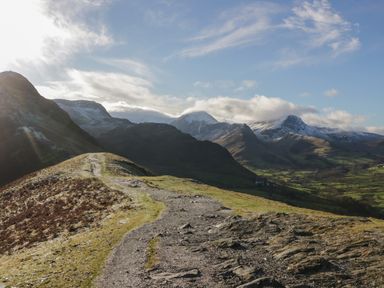 A mountain trail with grassy and rocky terrain and snow-capped peaks in the background