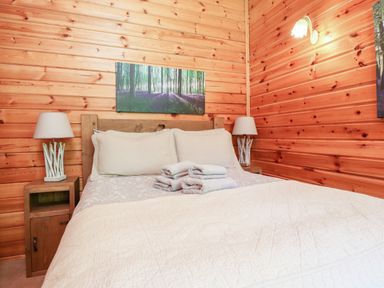 A bedroom with a wooden bed frame white pillows folded towels on bed wooden nightstands with lamps on each side and a forest painting on wooden walls at Gisburn Forest Lodge in Tosside near Wigglesworth