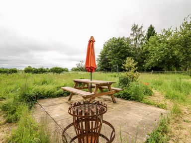 An outdoor seating area with a wooden picnic table and orange closed umbrella on a paved patio surrounded by grass and trees at Bluebell Cottage in Pembridge