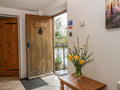 An entrance area with an open wooden door labeled Bluebell Cottage a wooden table with a glass vase of yellow flowers and a wall with a thermostat and a sign at Bluebell Cottage in Pembridge