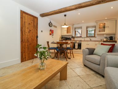 A living area with a wooden coffee table holding a vase with flowers a grey armchair with cushions and an open kitchen with dining table and chairs at Bluebell Cottage in Pembridge