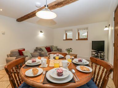 A dining table set with plates croissants jars of jam mugs and glasses of orange juice in a room with sofas and a television at Bluebell Cottage in Pembridge