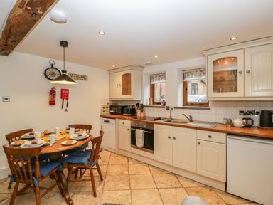 A kitchen with wooden cabinets a dining table set for breakfast and two windows at Bluebell Cottage in Pembridge