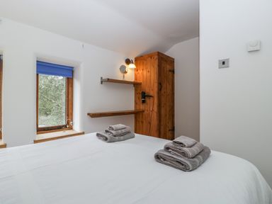 A bedroom with a bed dressed in white linens and folded towels on top a wooden wardrobe wall shelves and a window with a blue blind at Bluebell Cottage in Pembridge
