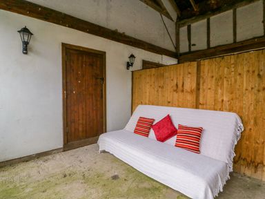 A covered outdoor seating area with a wooden door two wall lanterns and a white-covered sofa with three red cushions at Bluebell Cottage in Pembridge