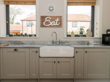 A kitchen sink area with a white apron front sink under two windows with wooden blinds and a wooden sign reading Eat above the sink at Pheasant's Roost Lelley near Burton Pidsea