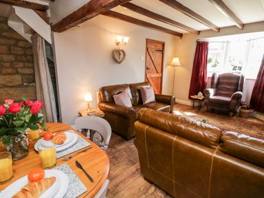 A living room with brown leather sofas a wooden door a wooden dining table with croissants and orange juice red roses and a plaid armchair by a window at The Lodge in Stainsacre near Whitby