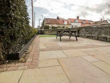 A paved outdoor patio area with a wooden picnic table and hedge bordering the side at The Lodge in Stainsacre near Whitby