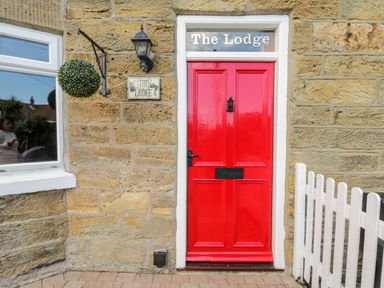 A red door with a black handle and mail slot on a stone wall with a hanging plant a lantern and a white picket fence at The Lodge in Stainsacre near Whitby