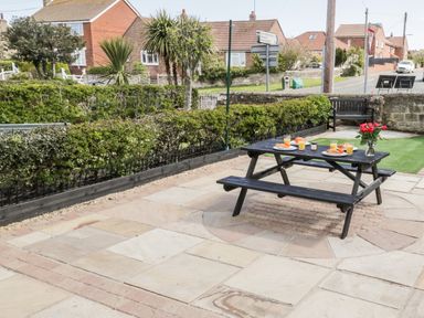 A patio with a wooden picnic table set with breakfast items and flowers near hedges and houses at The Lodge in Stainsacre near Whitby