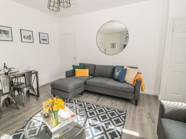 A living room with a gray sofa and colorful cushions a round mirror a glass coffee table with flowers and a dining table with metal chairs at Pebble Cove in Amble