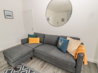 A grey sofa with colorful cushions and a blanket under a round mirror on the wall at Pebble Cove in Amble