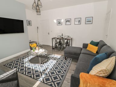 A living room with a gray sectional sofa with colored pillows a round glass coffee table on a patterned rug a dining table with four metal chairs and framed pictures on the wall at Pebble Cove in Amble
