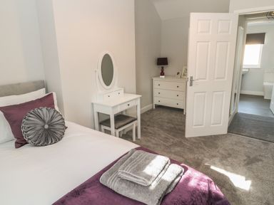 A bedroom with a bed, dressing table with mirror, chest of drawers, and open door leading to bathroom at Pebble Cove in Amble