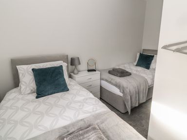 A bedroom with two single beds a white nightstand a lamp towels and teal pillows at Pebble Cove in Amble