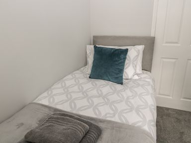 A single bed with patterned white bedding and a blue cushion with folded grey towels on top at Pebble Cove in Amble