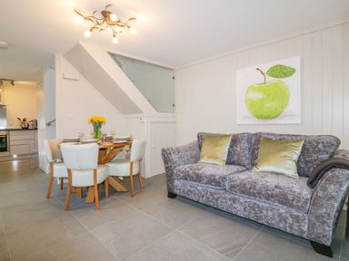 A dining area with a wooden table set for four and a sofa with cushions under a painting of a green apple at Apple Barn in Crantock