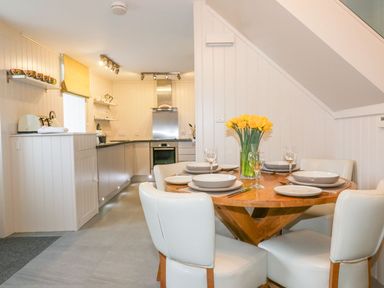 A dining area with a wooden round table set with plates glasses and a vase of yellow flowers next to a kitchen with cabinets and stainless steel appliances at Apple Barn in Crantock