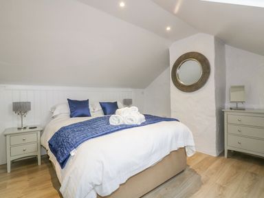 A bedroom with a bed with blue pillows and blanket a nightstand with a lamp a dresser with a mirror and a round wall mirror at Apple Barn in Crantock