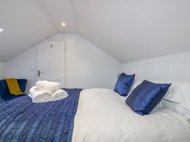 A bedroom with a bed dressed in white linens and blue pillows with towels on the bed and a blue chair with a yellow throw at Apple Barn in Crantock