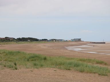 A sandy beach with grassy dunes and small colorful beach huts in the distance at 98 Cherry Park in Chapel St Leonards