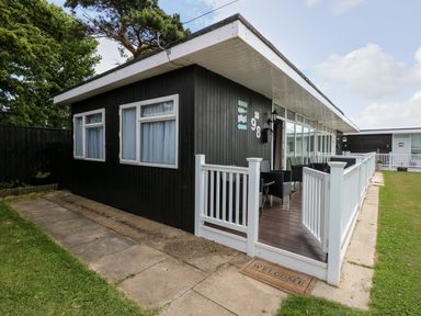 A black single-story house with white windows a white fenced porch with outdoor seating and a welcome mat at 98 Cherry Park in Chapel St Leonards