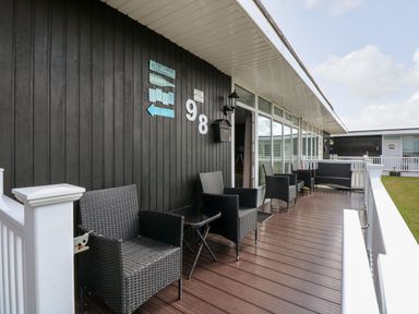 A wooden porch with black wicker chairs and a small table outside a building with dark walls at 98 Cherry Park in Chapel St Leonards