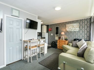 A small living area with a couch pillows a wooden chest of drawers a lamp a wall mirror a television two chairs and a kitchenette at 98 Cherry Park in Chapel St Leonards