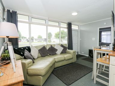 A living room with a leather sofa and pillows a wooden lamp table and a dining table with chairs at 98 Cherry Park in Chapel St Leonards