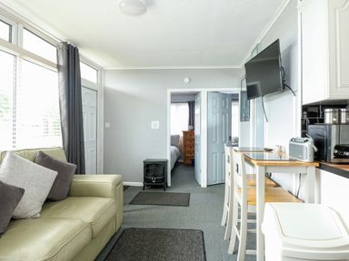 A living room with a sofa and cushions a small electric stove a wall mounted tv a high dining table with two chairs next to a kitchen area at 98 Cherry Park Chapel St Leonards