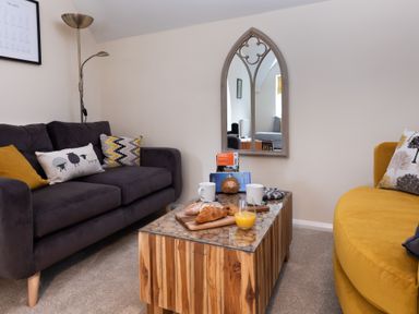 A living room with a dark gray sofa, yellow sofa, wooden coffee table with croissants and orange juice, and a gothic-style wall mirror at The Belfry in Windermere