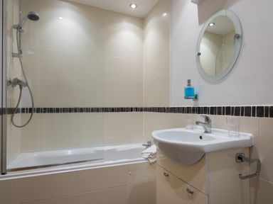 A bathroom with a bathtub a shower a sink with two glasses and a round mirror at The Belfry in Windermere