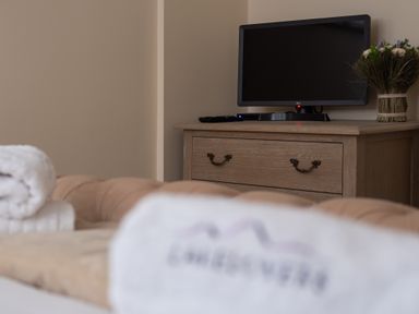 A TV on a wooden dresser with a remote and a flower arrangement on top in a bedroom at The Belfry in Windermere