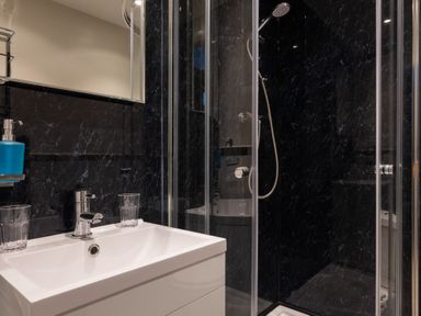 A bathroom with a white sink and faucet two glasses a blue soap dispenser a mirror and a glass shower enclosure with a handheld showerhead at The Belfry in Windermere