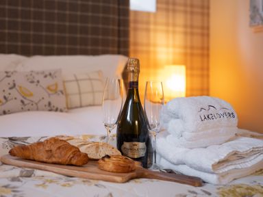 A bed with pillows a wooden board with croissant bread and pastry a bottle of sparkling wine two empty glasses and folded white towels at The Belfry in Windermere