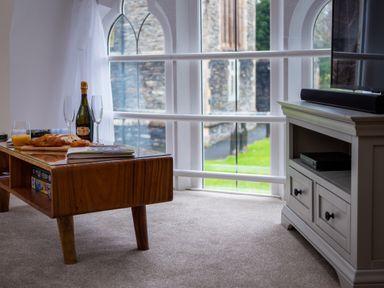 A wooden coffee table with croissants a bottle of champagne two glasses and a glass of orange juice by an arched window in a carpeted room at The Belfry in Windermere