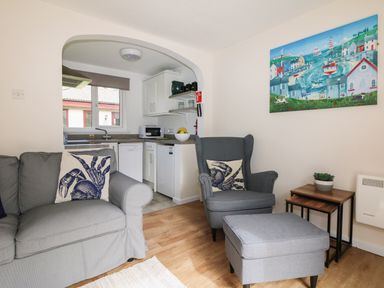 A living room with a gray armchair and ottoman next to nesting tables and a kitchen visible through an arch at Miles Retreat in Hayle