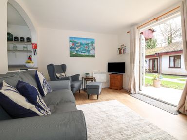 A living room with a grey sofa and armchair a small TV on a wooden stand and open doors to a garden at Miles Retreat in Hayle