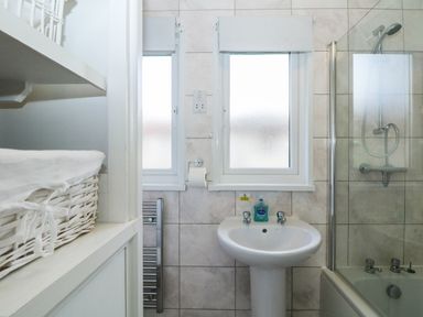 A bathroom with a pedestal sink soap bottle two frosted windows a shower over bathtub and a white wicker basket at Miles Retreat in Hayle