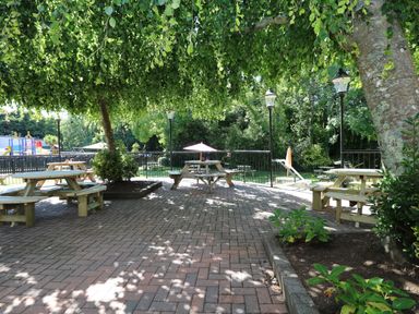 An outdoor seating area with wooden picnic tables under trees and a playground in the background at Miles Retreat in Hayle