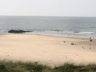 A sandy beach with people near the shore and grassy dunes in the foreground at Miles Retreat in Hayle