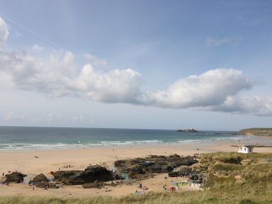 A sandy beach with rock formations and people near the ocean with a lighthouse on a small island in the background at Miles Retreat in Hayle