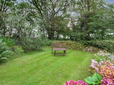 A garden area with a wooden bench and table on grass surrounded by trees and plants at The Roundhouse in Warbstow