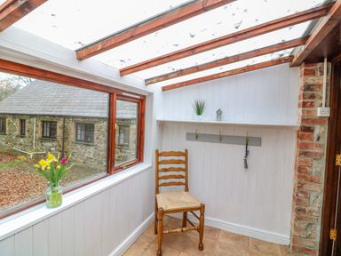 A small sunroom with a wooden chair a vase of flowers on the windowsill and a coat rack on the wall at The Roundhouse in Warbstow