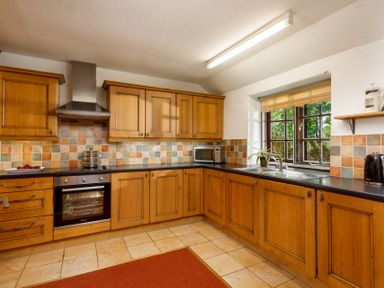 A kitchen with wooden cabinets a tiled backsplash an oven a microwave a sink and a window at The Roundhouse Warbstow