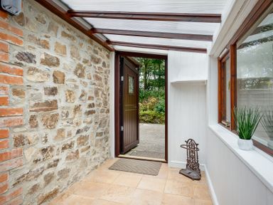 A hallway with a stone wall an open wooden door a window with a plant on the sill and a small umbrella stand at The Roundhouse in Warbstow