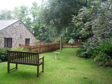 A garden with a wooden bench birdbath and a stone building surrounded by trees and flowers at The Roundhouse in Warbstow